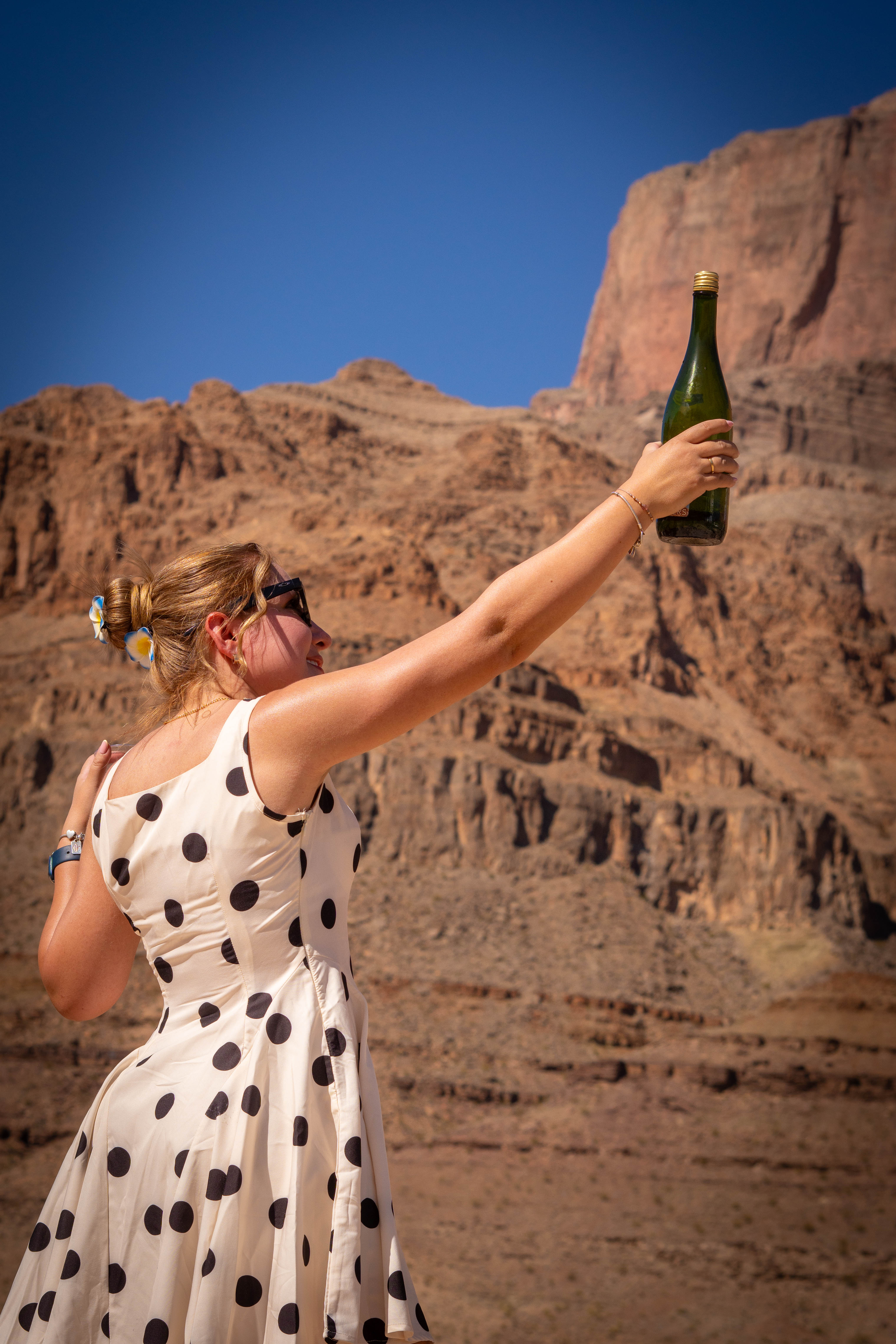 Mujer con vestido de lunares levantando una botella frente a las paredes rocosas del cañón